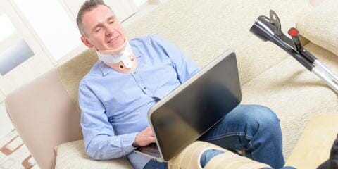 A person sitting on their couch at home with a neck brace and crutches, using their laptop to file a workers' compensation claim.