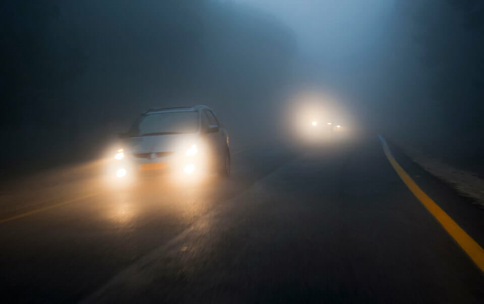 Several cars traveling in heavy fog down a two-lane road.